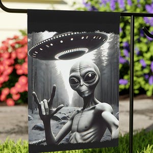 May include: Black and white garden flag depicting an alien with a UFO above, set against a forest scene. The alien is making a hand gesture. The flag is displayed in a garden, surrounded by greenery and flowers.