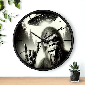 May include: Black and white clock featuring a Bigfoot with a tongue sticking out, giving a rock and roll hand gesture. A UFO is flying above Bigfoot, with trees in the background.