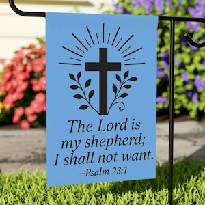 May include: A light blue garden flag with a black cross design, sunburst rays, and leafy branches. The text reads "The Lord is my shepherd; I shall not want. -Psalm 23:1". The flag is displayed on a black metal stand in a garden setting.