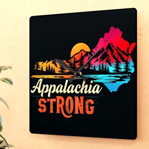 Op de afbeelding: Vierkante wandklok met een zwarte wijzerplaat met een kleurrijk berg- en meerlandschap met een zonsondergang. De klok leest "Appalachia STRONG" in witte en oranje tekst. De wijzers van de klok zijn zwart.
