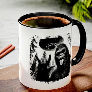 May include: White ceramic mug with a black handle and rim. The mug has a black and white graphic of a Bigfoot creature, a UFO, and a hand making a rock and roll gesture. The mug is filled with coffee.