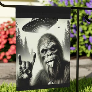 May include: A black and white garden flag featuring a Bigfoot with its tongue sticking out, giving the rock and roll hand gesture. A UFO is flying above Bigfoot in the background.