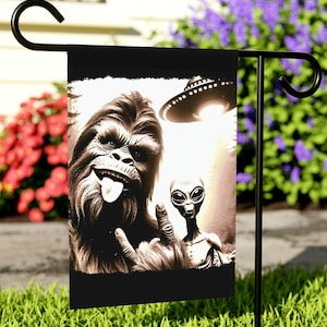 May include: A black garden flag with a sepia-toned image of Bigfoot sticking out its tongue and giving a peace sign, alongside a grey alien. A UFO hovers above them. The flag is displayed on a black metal stand.