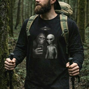May include: Black long-sleeved shirt with a graphic of a smiling Bigfoot and an alien taking a selfie under a UFO. The shirt is worn by a person in a forest setting, wearing a black cap and carrying a backpack.