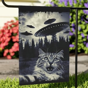 May include: A monochrome garden flag showing a startled cat with a forest backdrop and flying saucers in the sky. The flag is displayed on a black metal stand in a garden with grass and flowers.