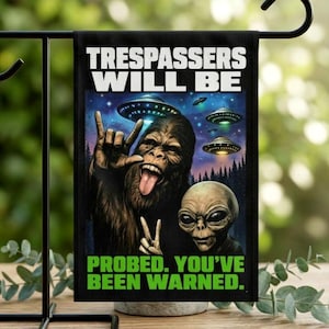 May include: A rectangular garden flag featuring a brown Bigfoot making a rock-and-roll hand gesture and a grey alien making a peace sign. Above them are flying saucers in a night sky. The text reads: TRESPASSERS WILL BE PROBED. YOU'VE BEEN WARNED.