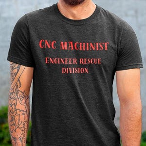 May include: A dark gray t-shirt with the text "CNC Machinist Engineer Rescue Division" printed in red.