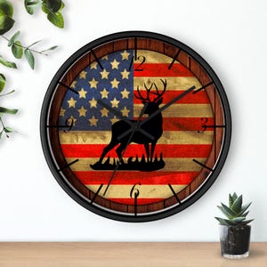 May include: A round wall clock with a black frame and a distressed American flag design. A silhouette of a deer is in the center of the flag. The clock face has Roman numeral markings.