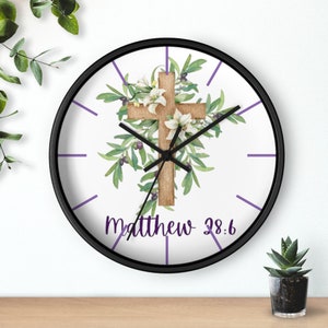Könnte beinhalten: Eine weiße runde Uhr mit schwarzem Rahmen und violetten Stundenmarkierungen. Das Zifferblatt der Uhr zeigt ein Holzkreuz mit Olivenzweigen und weißen Lilien. Der Text "Matthäus 28:6" ist unter dem Kreuz gedruckt.