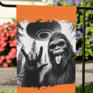 May include: Orange garden flag with a black and white image of a Bigfoot-like creature with its tongue out and a hand making a rock and roll gesture. A UFO hovers above the creature. The background includes a forest scene.