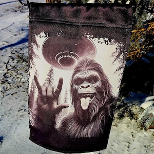 Puede incluir: Una bandera de jardín en blanco y negro con un gráfico de un Bigfoot haciendo el signo de la mano del rock and roll con un OVNI en el fondo.
