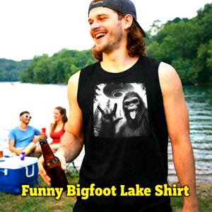May include: Black tank top with a graphic of Bigfoot giving a rock-on hand gesture and a UFO. The text "Funny Bigfoot Lake Shirt" is displayed in yellow. A person is wearing the tank top and holding a beer bottle.