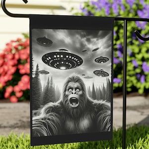 May include: Black and white garden flag depicting a startled Bigfoot with a forest background and flying saucers in the sky. The flag hangs on a black metal stand, with green grass and colourful flowers surrounding it.