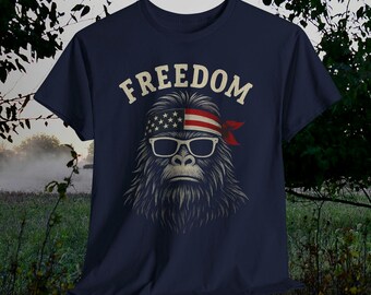 Puede incluir: Camiseta azul marino con un gráfico de un Bigfoot con gafas de sol y una cinta para la cabeza con el diseño de la bandera estadounidense. La palabra "FREEDOM" está impresa en blanco encima del gráfico. La camiseta es de cuello redondo.