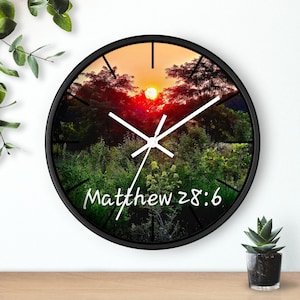 Op de afbeelding: Een ronde wandklok met een zwart frame. De wijzerplaat toont een zonsondergang met bomen en groen. De tekst "Matthew 28:6" staat in witte letters onderaan.