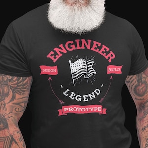 Puede incluir: Una camiseta negra con un diseño gráfico rojo y blanco que dice "Engineer" con una bandera estadounidense y las palabras "Design", "Build", "Legend" y "Prototype".
