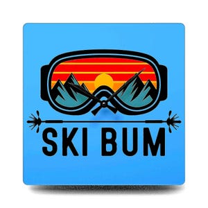 May include: A blue square clock with a ski goggle design. The goggle lens has a mountain and sunset scene. The clock face is hidden behind the goggle lens. The text "SKI BUM" is printed below the goggle.