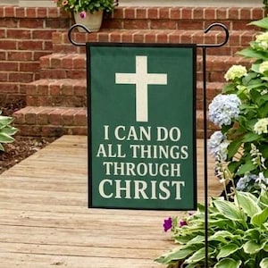 Op de afbeelding: Een groene tuinvaandel met een wit kruis en de tekst "I CAN DO ALL THINGS THROUGH CHRIST". De vlag wordt buiten weergegeven, met een houten loopbrug en groen in de achtergrond.