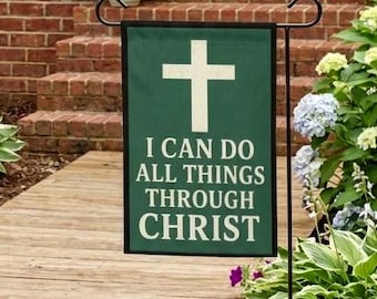 Puede incluir: Una bandera de jardín verde con una cruz blanca y el texto "I CAN DO ALL THINGS THROUGH CHRIST". La bandera se muestra al aire libre, con una pasarela de madera y vegetación en el fondo.