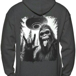 May include: A dark gray hooded sweatshirt with a black and white graphic print of a Bigfoot giving the 'I love you' hand sign with a UFO flying overhead.