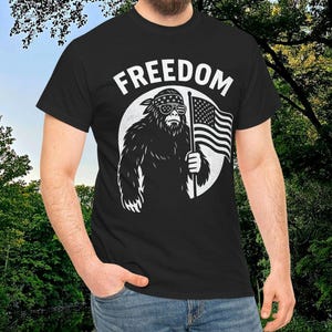 以下が含まれることがあります： 「FREEDOM」の文字と、サングラスとバンダナをつけたビッグフットがアメリカ国旗を持っている白いグラフィックがプリントされた黒いTシャツ。柔らかい素材で作られています。