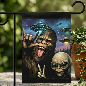 May include: A garden flag featuring a Bigfoot character and an alien making hand gestures with flying saucers in the background. The flag has a black border and is displayed outdoors.
