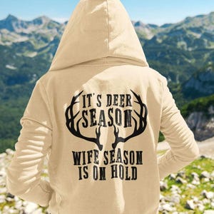 May include: A beige hoodie with a black deer antler graphic and the text "It's Deer Season Wife Season Is On Hold".