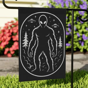 May include: Black garden flag with a white illustration of an alien figure, UFOs, trees, and stars within an oval frame. The flag is mounted on a black metal garden stake, set against green grass and vibrant flowers. A decorative item for the garden.