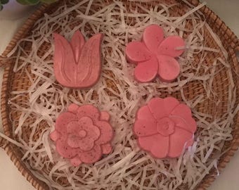 May include: A woven basket with handles holds four pink floral-shaped items nestled on white shredded paper. The flowers include a tulip, a five-petal flower, and two other detailed floral designs. The basket is brown and sits on a white surface.