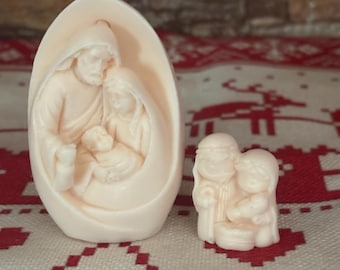 May include: Two small, off-white nativity figurines. One is an oval-shaped sculpture depicting Mary, Joseph, and baby Jesus. The other is a smaller, rectangular figurine of the holy family. The figurines are set against a red and white patterned background.