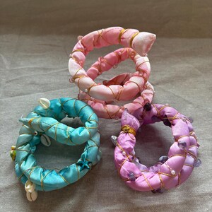 Puede incluir: Tres servilleteros decorativos en tonos rosa, azul y morado. Los anillos están hechos de tela y adornados con alambre dorado, cuentas y conchas marinas. Los anillos rosas tienen perlas, el anillo azul tiene conchas y una estrella de mar, y el anillo morado tiene cuentas moradas.