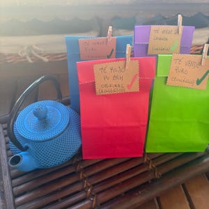 May include: A bright blue cast iron teapot with a textured surface sits beside four colourful paper bags in blue, red, purple, and green. Each bag is sealed with a wooden clip holding a brown paper tag with handwritten text and a checkmark.