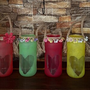 May include: Six colorful glass jars with heart cutouts and rope handles. Each jar is painted a different color: blue, red, green, red, yellow, and green. The jars are decorated with flowers around the rim, creating a decorative and charming display.