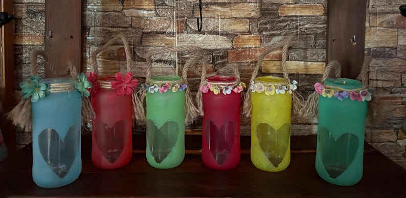 May include: Six colorful glass jars with heart cutouts and rope handles. Each jar is painted a different color: blue, red, green, red, yellow, and green. The jars are decorated with flowers around the rim, creating a decorative and charming display.