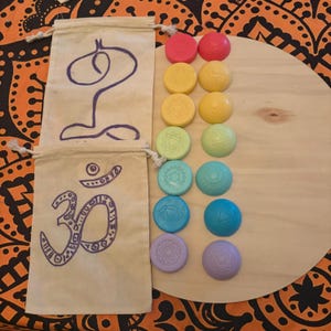 Puede incluir: Una colección de piedras de chakra coloridas dispuestas sobre una superficie de madera, junto con dos bolsas beige con cordón y ilustraciones moradas. Las piedras son en tonos de rojo, amarillo, verde, azul y morado, cada una con un símbolo de chakra.