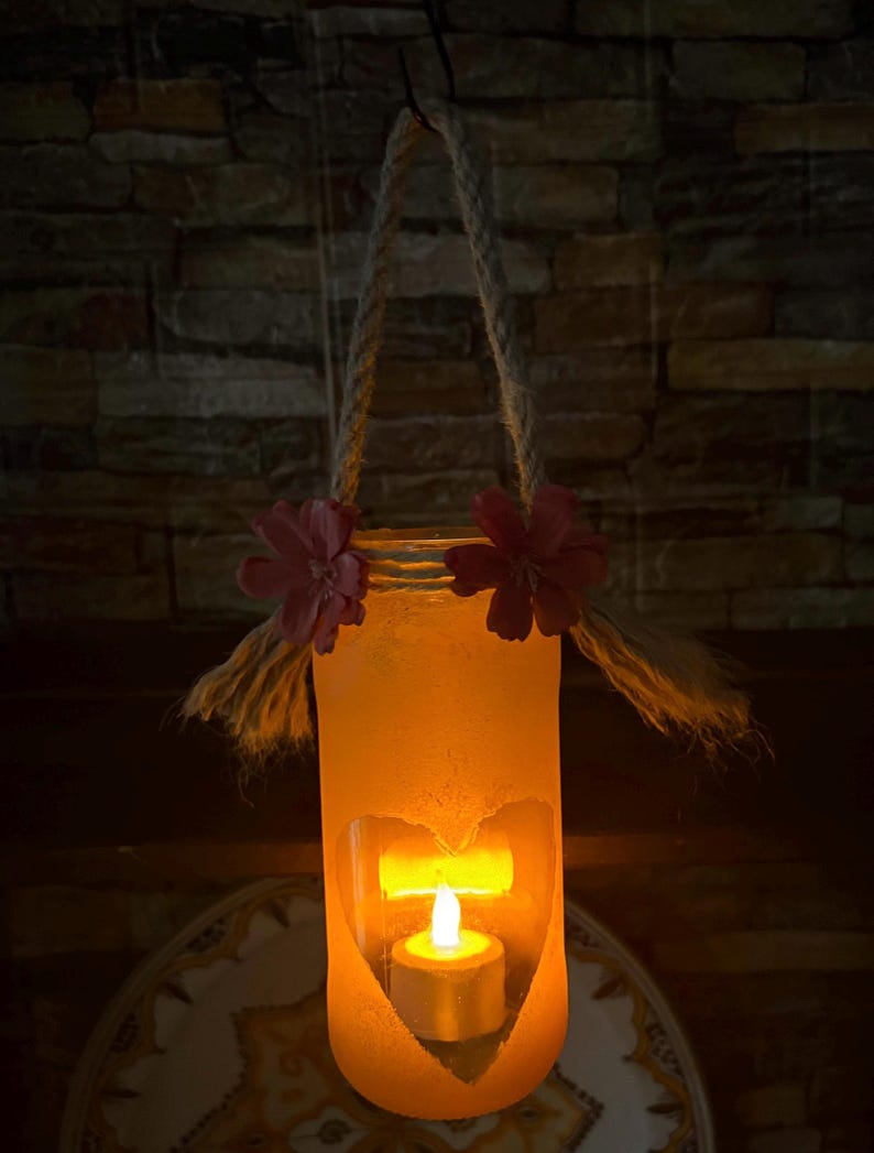 May include: A hanging candle holder with a heart-shaped cutout, illuminated by a small candle. The holder is cylindrical, with a rope handle and adorned with pink flowers. The warm glow creates a cozy ambiance.
