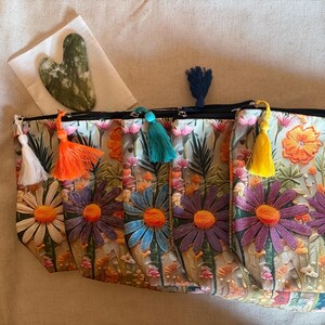 Puede incluir: Cuatro neceseres con estampado floral y tiradores de cremallera con borlas de colores. Las bolsas presentan un estampado de margaritas con flores naranjas, moradas y azules. Una herramienta de gua sha de jade verde descansa sobre un paño blanco.