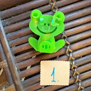Puede incluir: Un objeto verde brillante y translúcido con forma de rana, con dos ojos prominentes, una boca ancha sonriente y una barriga texturizada. Debajo de la rana hay un cuadrado de papel color canela con bordes dentados y el número 1 escrito en azul.