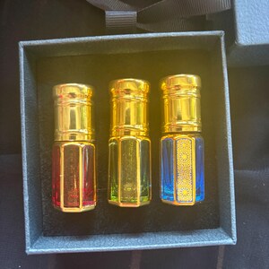 Può includere: Tre piccole boccette di profumo in vetro con tappi dorati disposte in una scatola grigio scuro. Le boccette sono rossa, verde e blu, con quella blu che presenta un motivo geometrico dorato sul lato. Ogni boccetta ha una base in vetro esagonale.