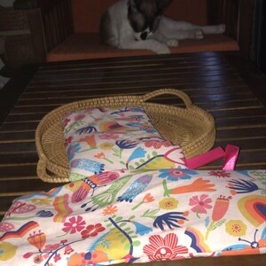 Puede incluir: Un tejido blanco con un colorido estampado floral y de pájaros, apoyado sobre una mesa de madera. Una cesta tejida contiene un artículo a juego. Se adjunta una cinta rosa. Un gato descansa en una silla en el fondo.