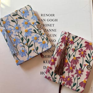 Puede incluir: Dos cuadernos bordados con diseños florales. Uno presenta flores azules, el otro rosas. Ambos tienen borlas y cierres de botón. El texto "RENOIR, VAN GOGH, MONET" es visible en el fondo.