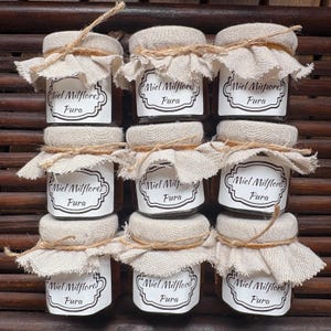 Pode incluir: Nove pequenos frascos de vidro cheios de mel, cada um coberto com um tecido de juta bege preso por um laço de corda castanho. Cada frasco apresenta um rótulo retangular branco com uma borda preta decorativa e o texto Miel Milflores Pura.