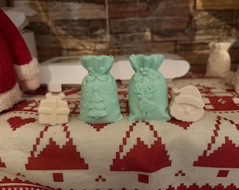 May include: A festive arrangement of Christmas-themed soaps. Two light green gift bag soaps with embossed designs of a Christmas tree and a reindeer. A white gift-shaped soap and a Santa Claus face soap are also present.