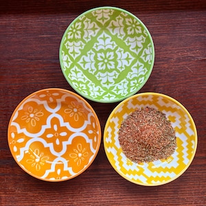 Op de afbeelding: Drie kleine ronde keramische schaaltjes met verschillende patronen. Eén schaaltje is limoengroen met een wit bloemmotief, één is oranje met een wit geometrisch bloemendesign en één is geel met een wit zigzagpatroon gevuld met droge kruiden.