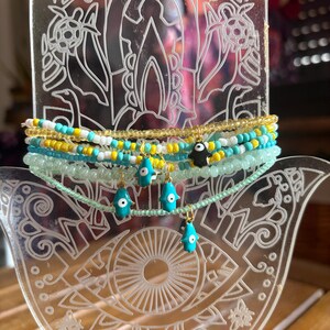 Puede incluir: Una colección de pulseras y collares de cuentas en tonos turquesa, amarillo, blanco y dorado. Algunas pulseras presentan pequeños dijes, incluyendo un pájaro negro y colgantes de mano de hamsa turquesa. La joyería se exhibe en un soporte decorativo transparente.
