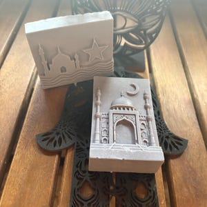 Puede incluir: Dos pastillas de jabón rectangulares de color gris con diseños en relieve. Una muestra la silueta de una mezquita con una estrella y líneas onduladas. La otra muestra una mezquita detallada con cúpula central, minaretes y una luna creciente.