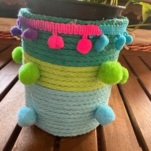 Puede incluir: Una maceta decorativa envuelta en cuerda y adornada con detalles coloridos. La maceta presenta capas de cuerda turquesa, verde y verde lima, acentuada con pompones y un ribete rosa. Contiene una planta verde.