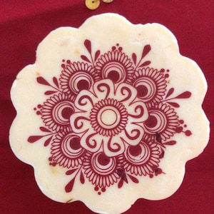 Puede incluir: Un posavasos crema con borde festoneado y un detallado diseño floral burdeos. El posavasos presenta un motivo circular central rodeado de pétalos y espirales estilizados. Tres pequeños adornos dorados se encuentran en la esquina superior izquierda sobre un fondo rojo.