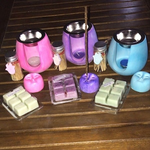 Puede incluir: Una colección de artículos de aromaterapia, que incluyen difusores de aceites esenciales rosas, morados y azules con cuencos plateados. También se muestran pequeños frascos de cerillas, ceras perfumadas y velas con forma de cara en varios colores. Los artículos están dispuestos sobre una superficie de madera.