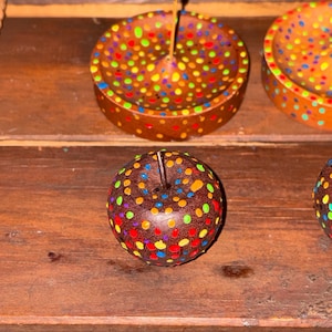 Puede incluir: Porta inciensos de madera pintados a mano y una manzana decorativa. Los porta inciensos son marrones con lunares coloridos. La manzana es marrón con lunares coloridos y un tallo de madera. Los artículos se exhiben sobre una superficie de madera.